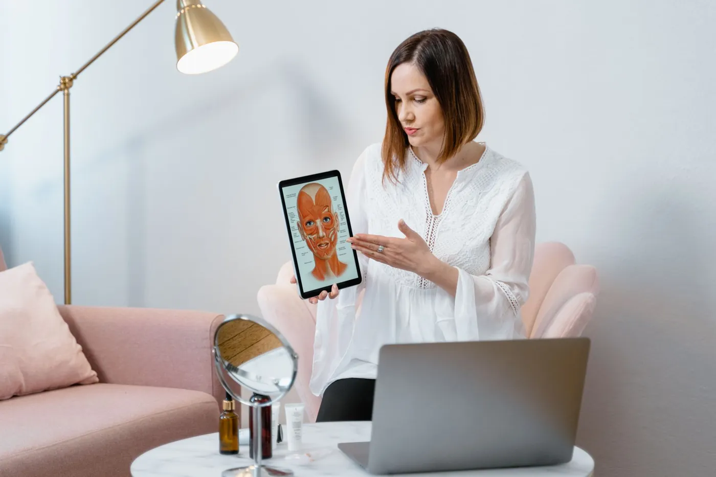 Aesthetic provider in a white blouse walking a client through a treatment plan on a tablet in a chic consultation lounge with a brass lamp