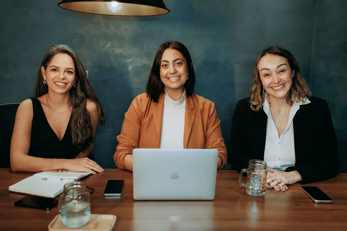 Three smiling professional women at a meeting table with a laptop