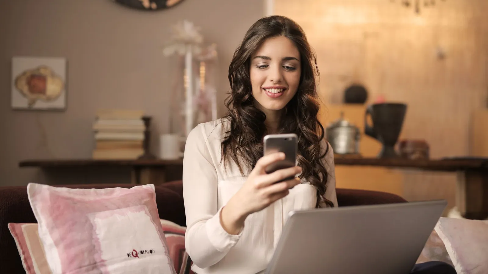 Woman smiling at her smartphone with a laptop open in a warmly lit, refined living space