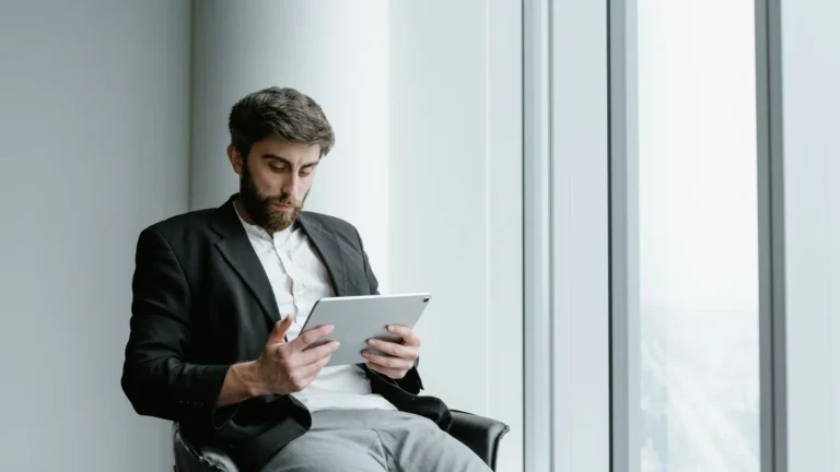 Man in a dark suit working on a tablet while seated by a large office window