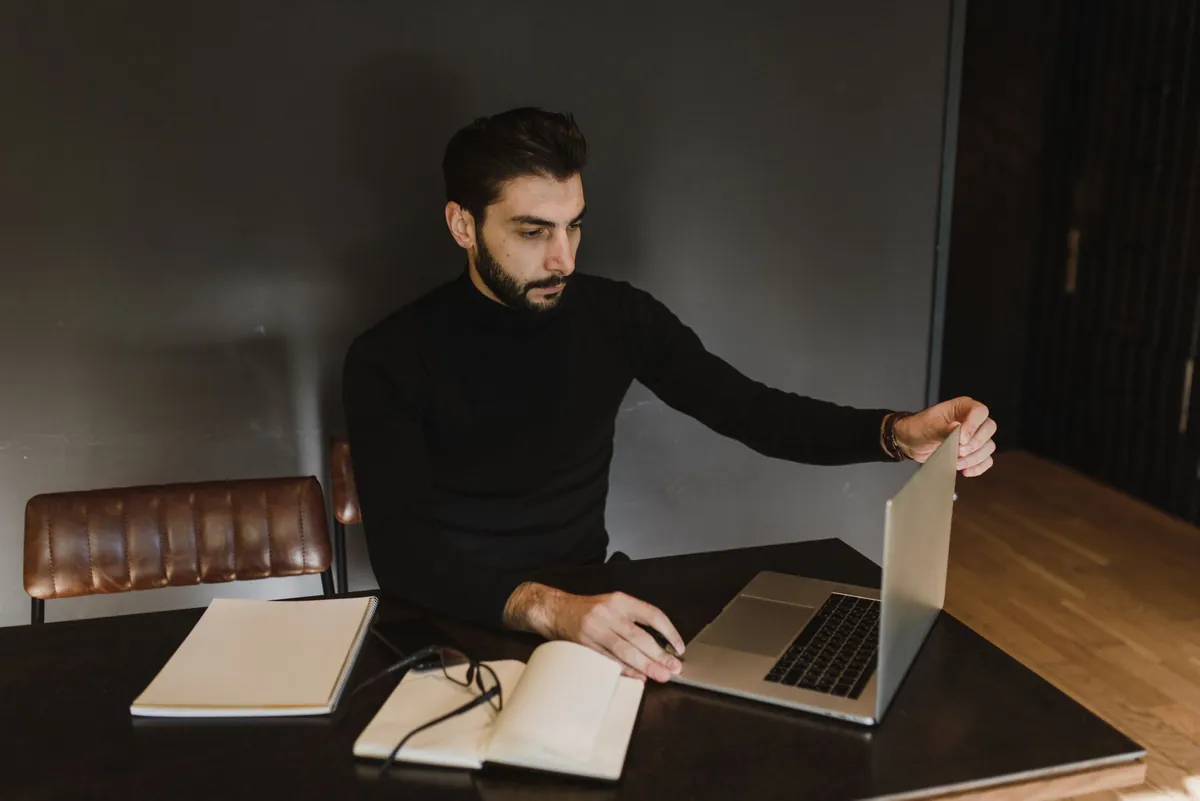 Man in a black sweater opening a laptop at a dark wood desk