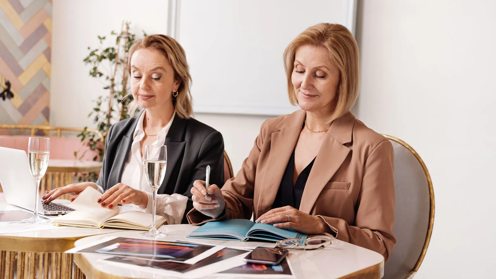 Two practice leaders in tailored blazers reviewing listings and notes at a brass café table