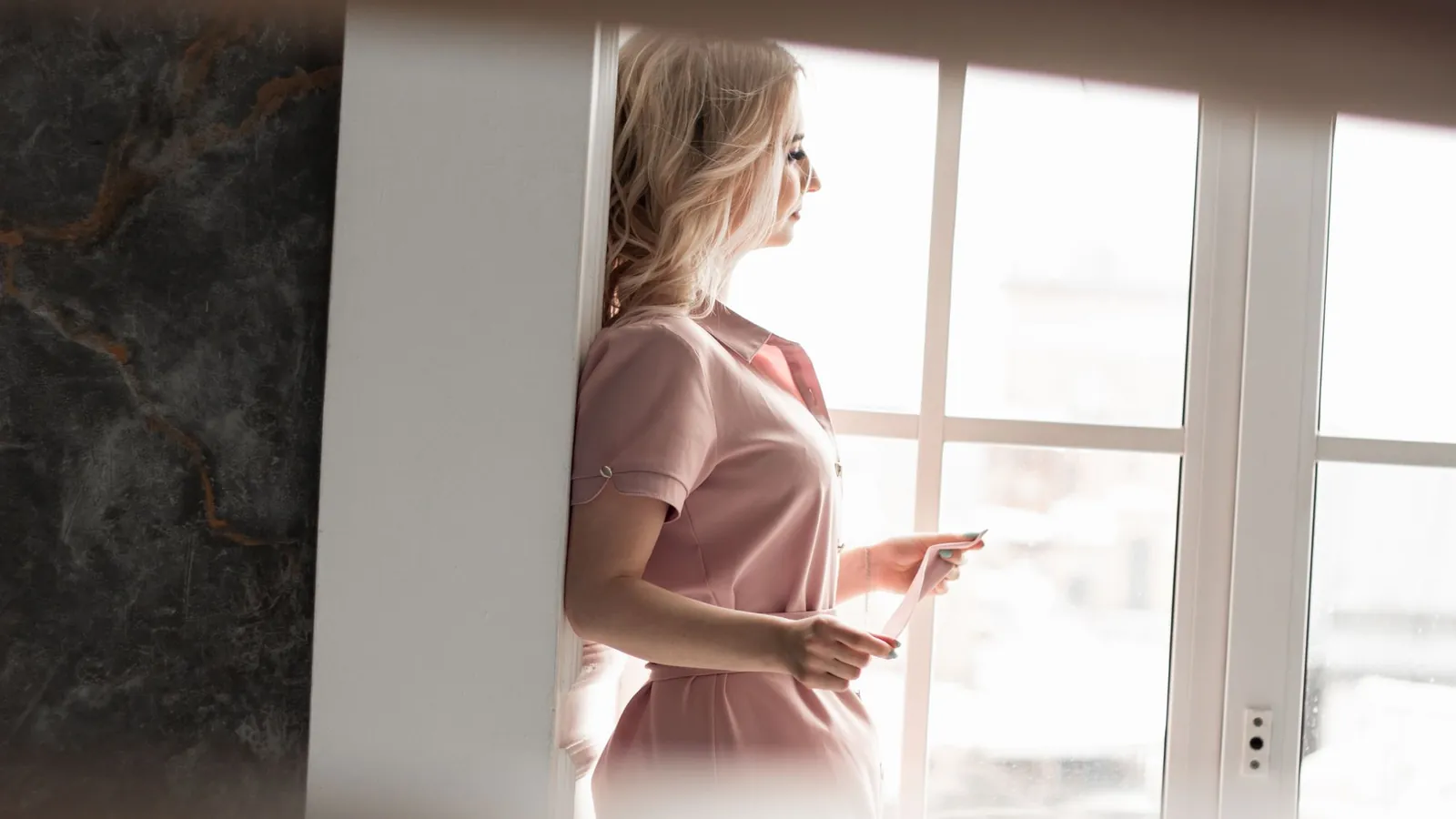 Woman in a blush blouse reading notes by a tall window in soft natural light