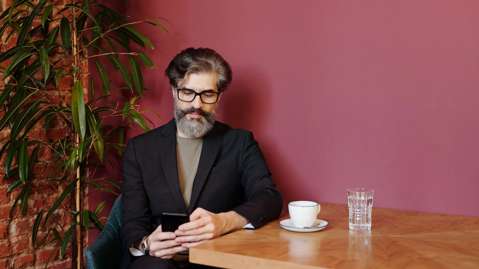 Stylish bearded man in a dark blazer using his smartphone at a cafe table against a red wall