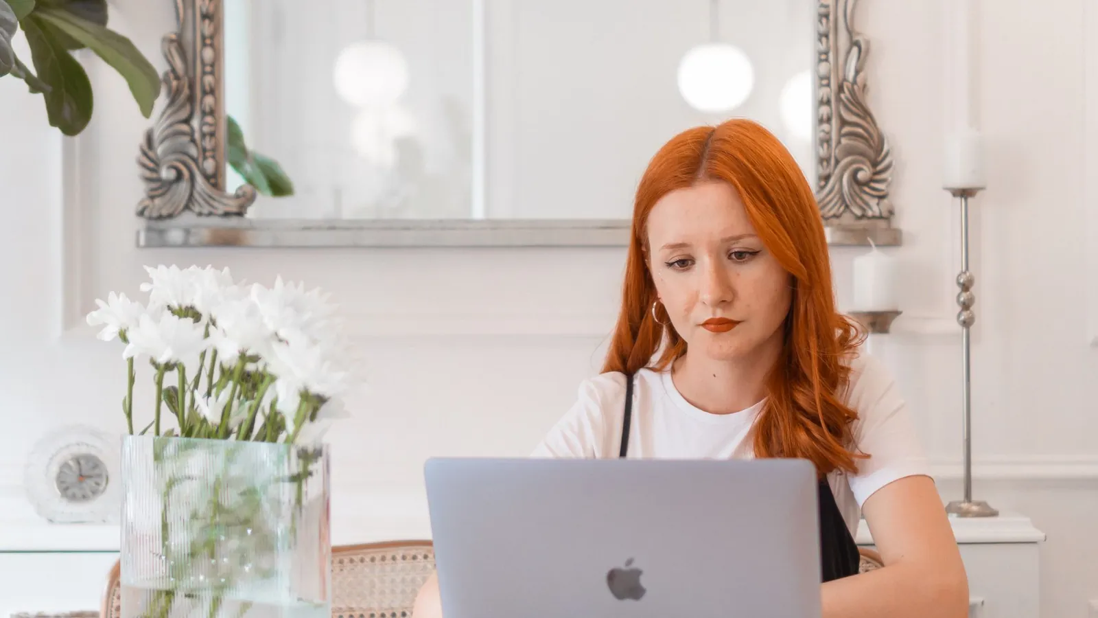 Woman working on a laptop at a table with white flowers and an ornate silver mirror behind her