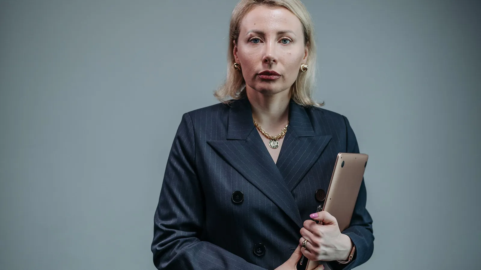 Confident blonde owner in a navy blazer holding a tablet against a soft gray wall