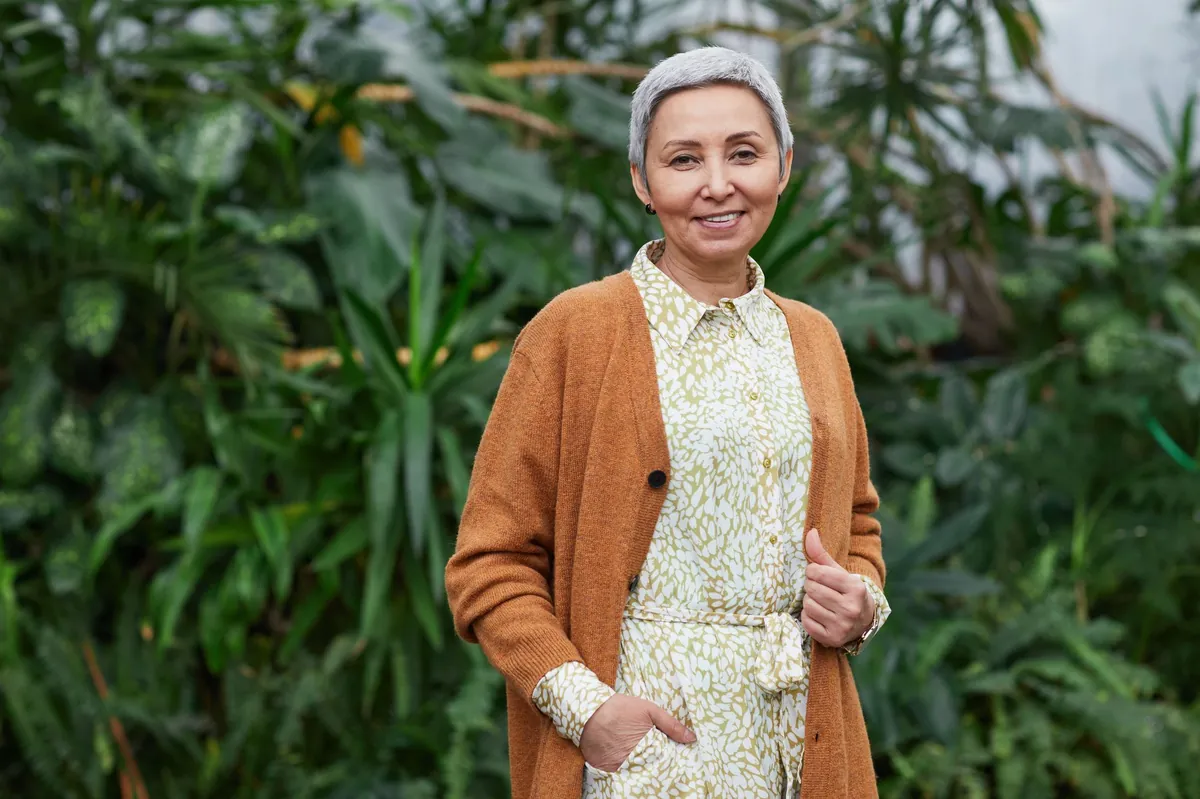 Joyful senior woman in a stylish cardigan smiling beside lush green plants