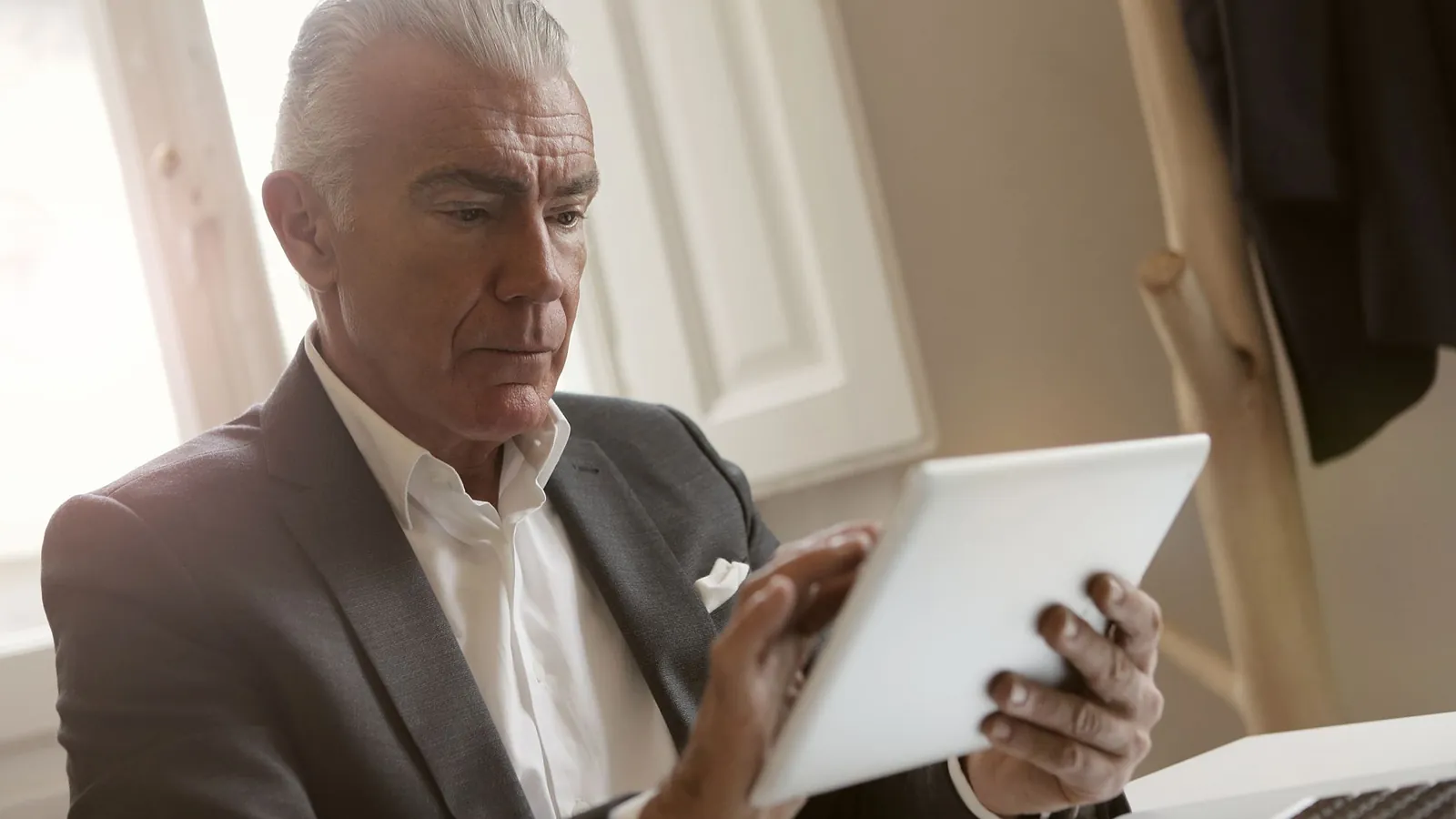 Senior businessman in a gray suit reviewing results on a tablet beside a large window