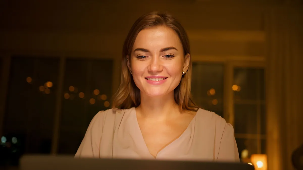 Smiling woman lit by a laptop screen in a softly lit evening room