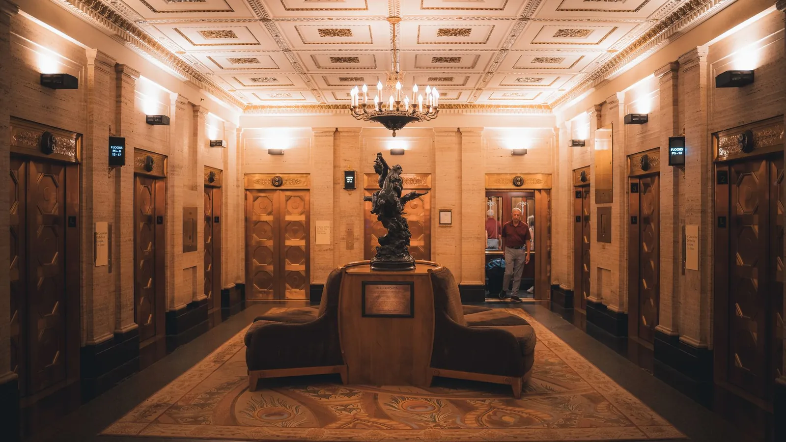 Grand gilded lobby with paneled walls, a chandelier and a central bronze statue