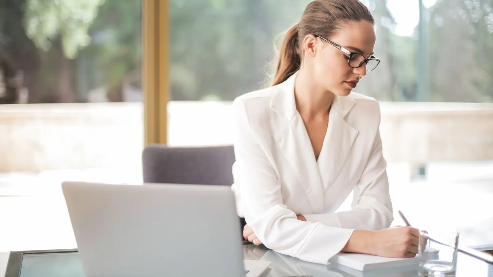 Professional in a white blouse writing notes at a desk beside a laptop