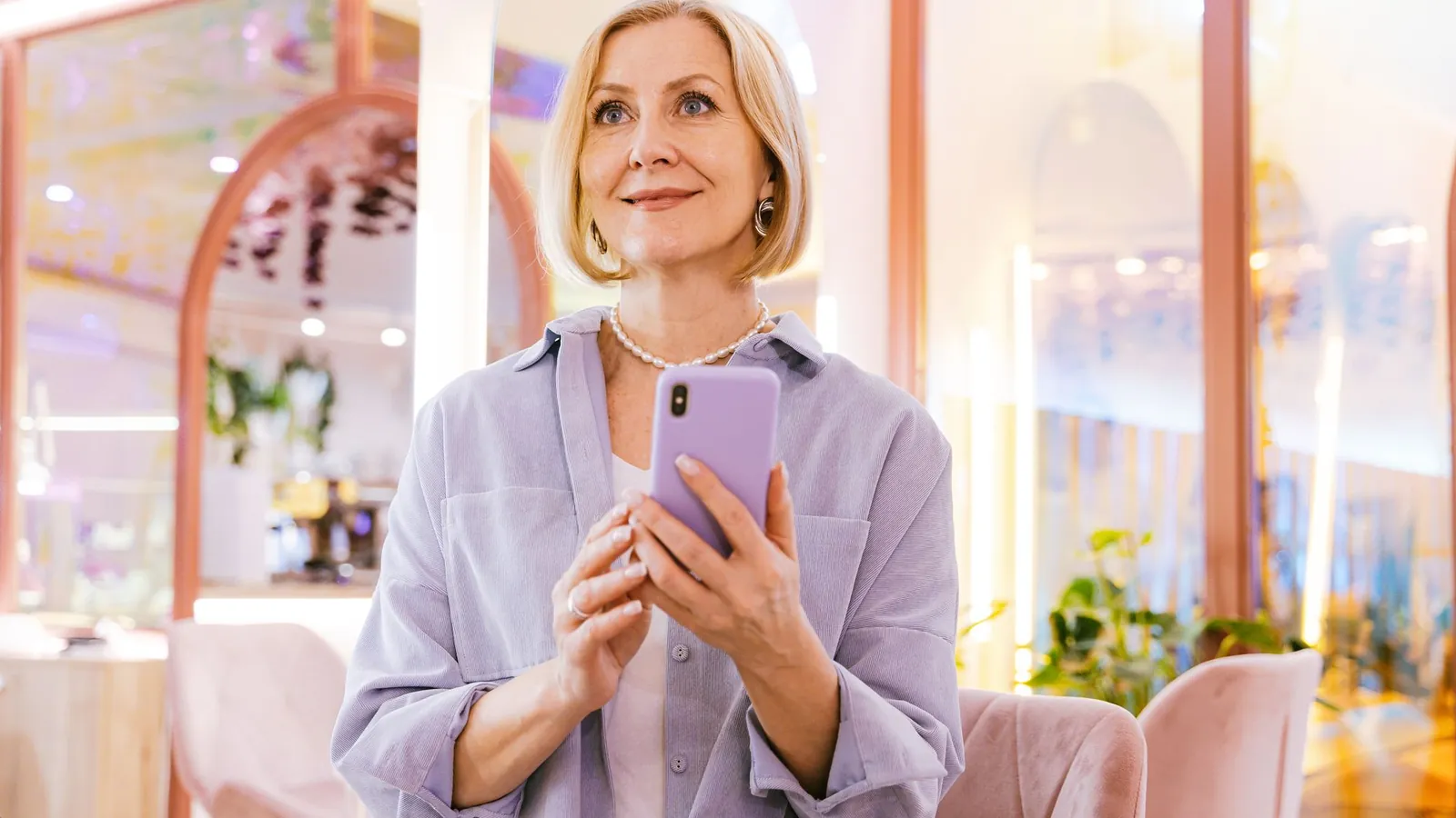 Smiling woman in a lavender blouse holding a current smartphone in a bright, rose-toned interior