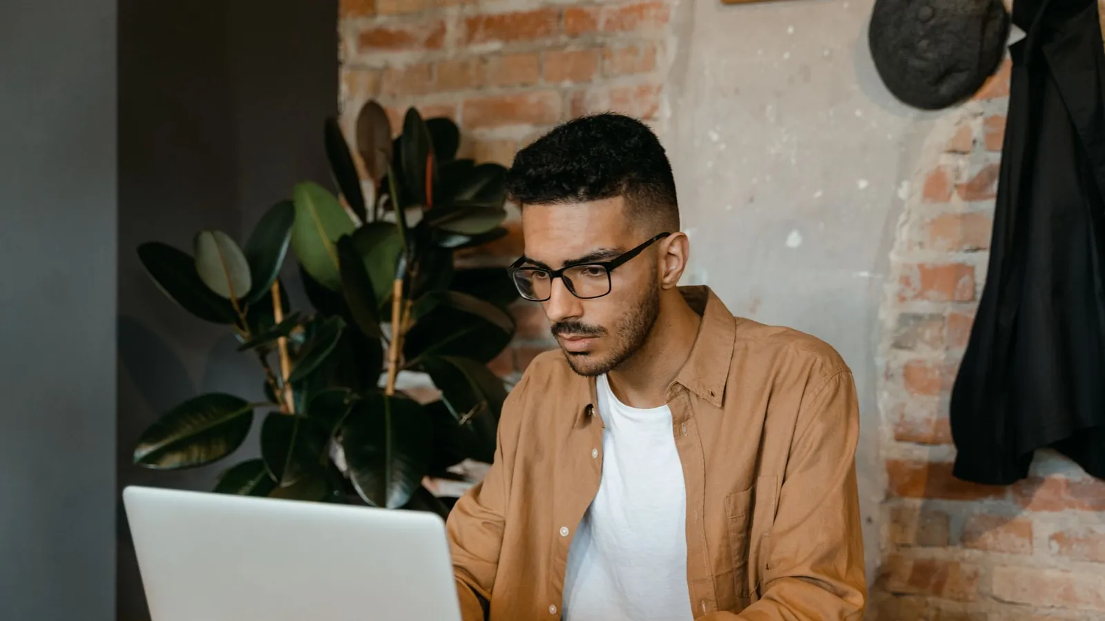 Practice owner in glasses working on a laptop in a cafe with brick walls and plants