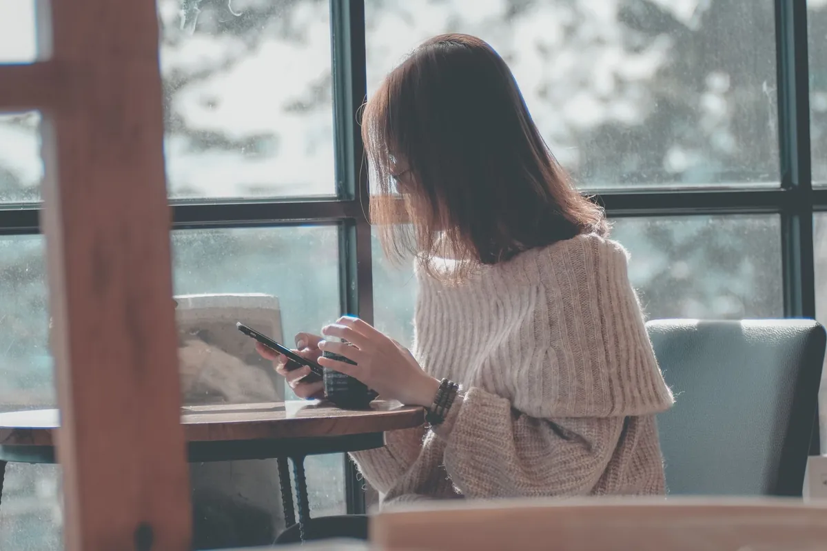 Woman scrolling her phone at a cafe table beside a window
