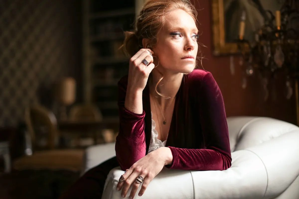 Woman in a burgundy velvet top seated thoughtfully in a dim, elegant room