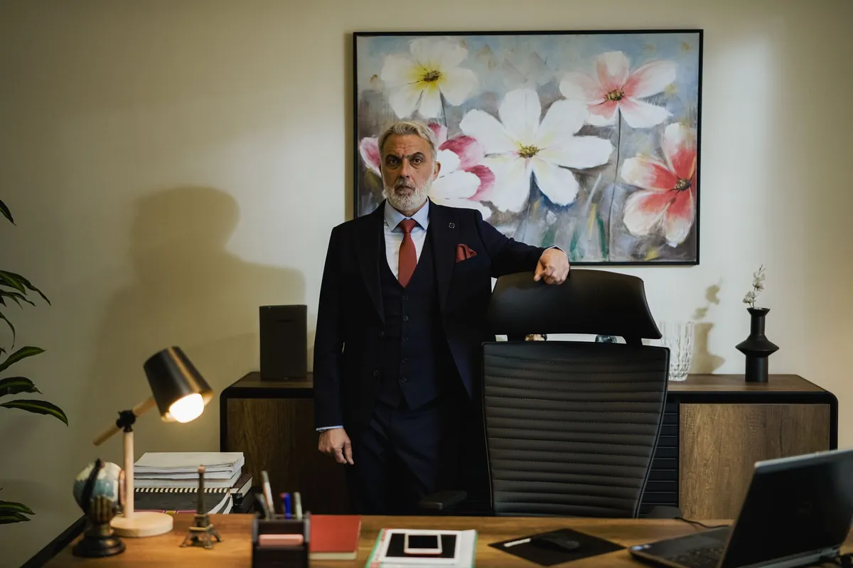 Executive in a dark suit standing behind his desk beneath a floral painting