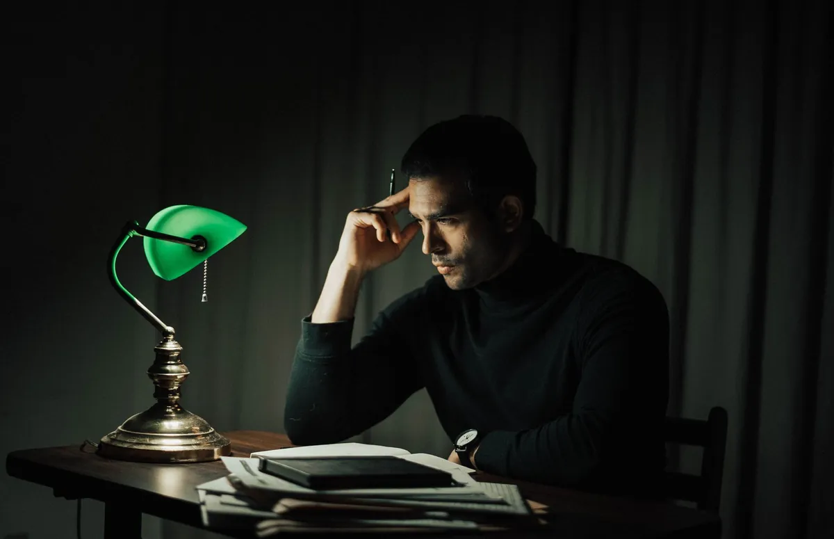 Man reading late at a dark desk beside a green banker lamp