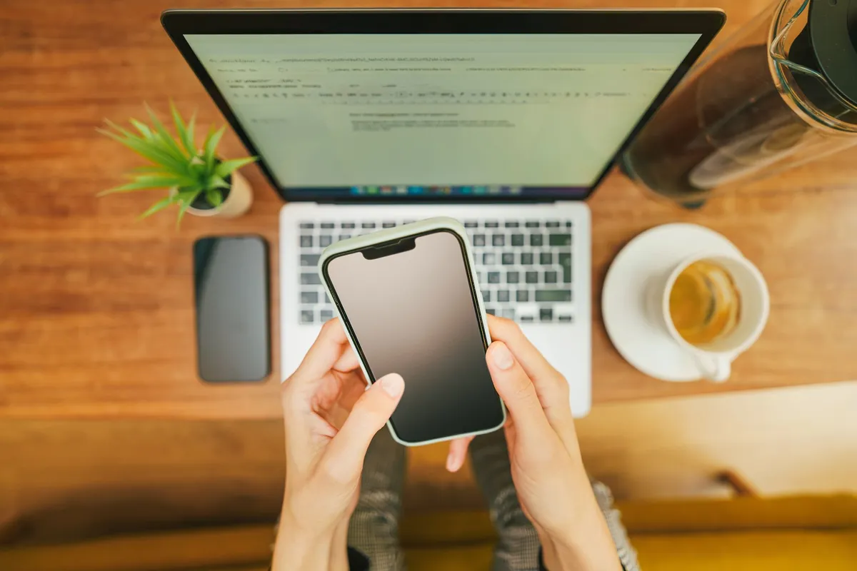 Hands holding a smartphone over a laptop with coffee in a home office