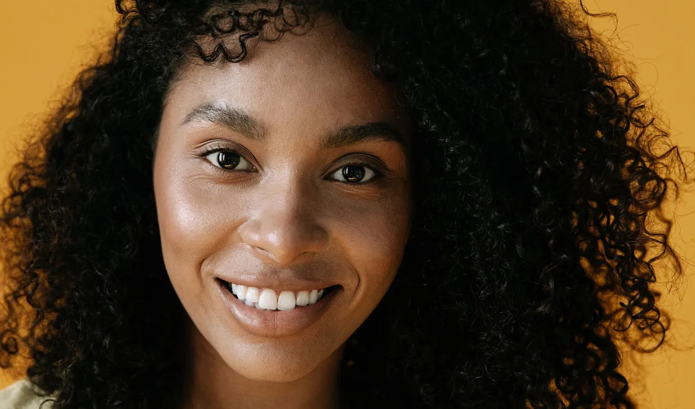 Smiling aesthetic client with radiant skin and natural curls against a warm golden backdrop