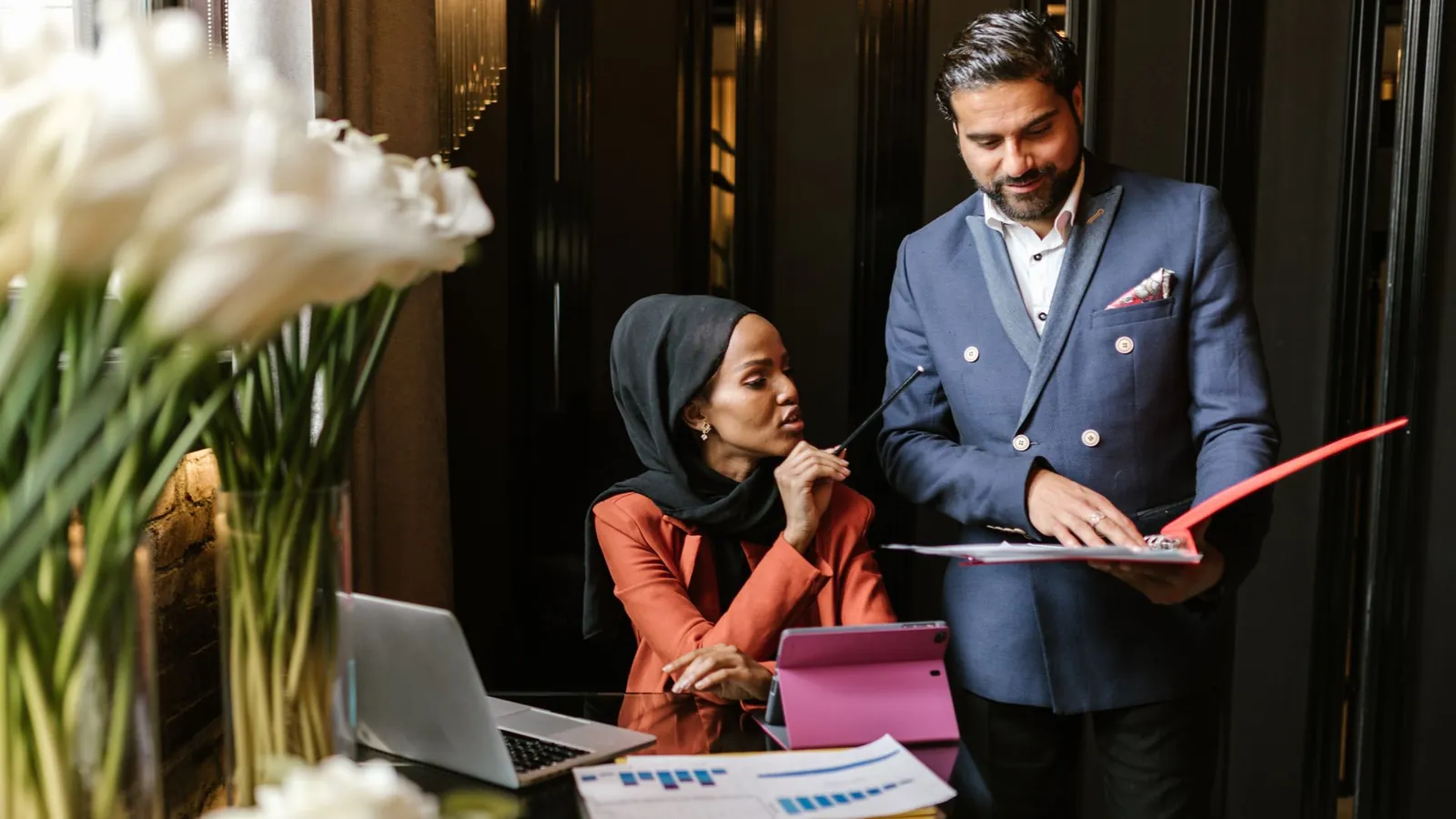 Marketing strategist in a tailored navy blazer reviewing a growth report with a practice owner at a glass desk with white calla lilies, warm light