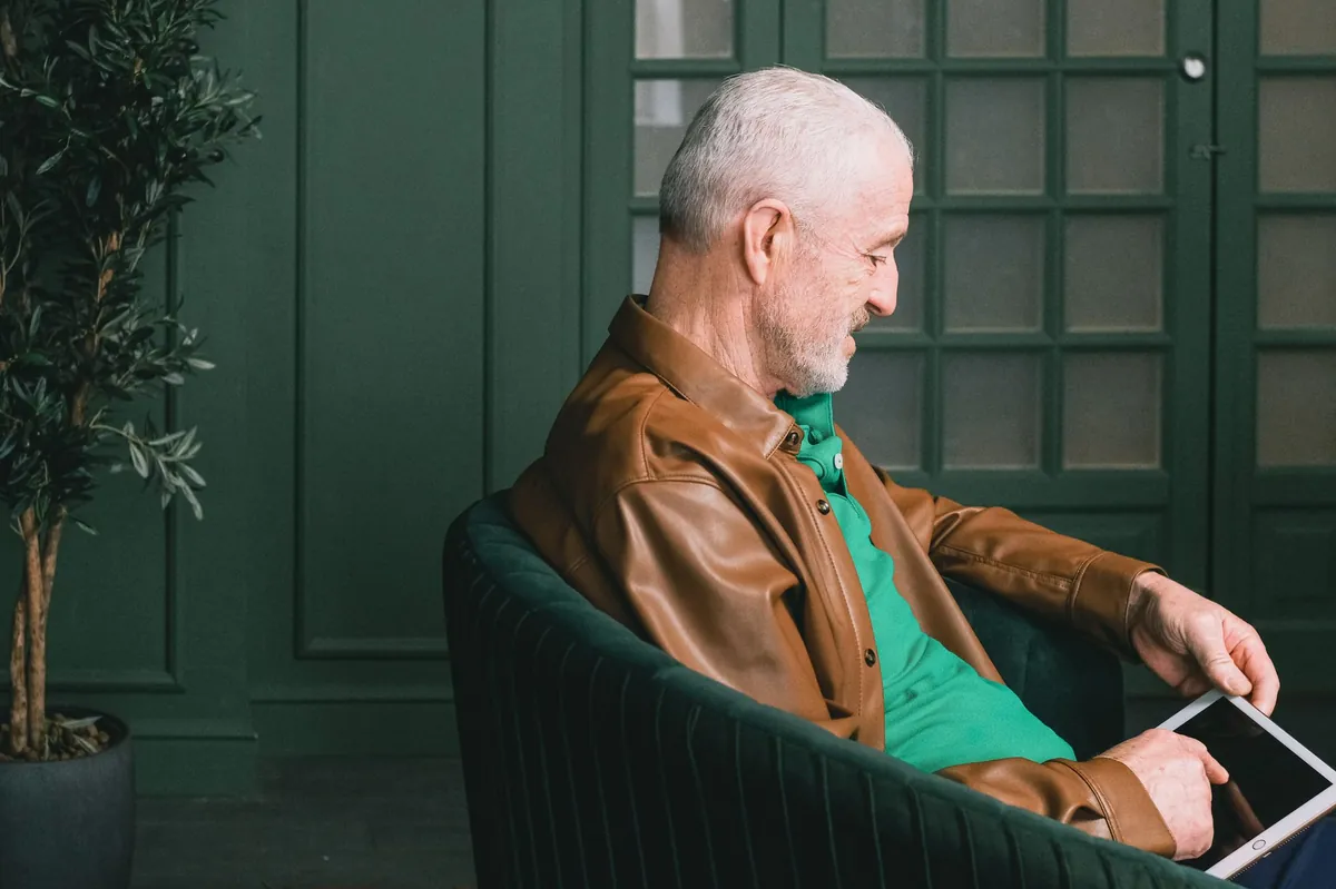 Silver-haired man in a leather jacket reading a tablet in a green armchair