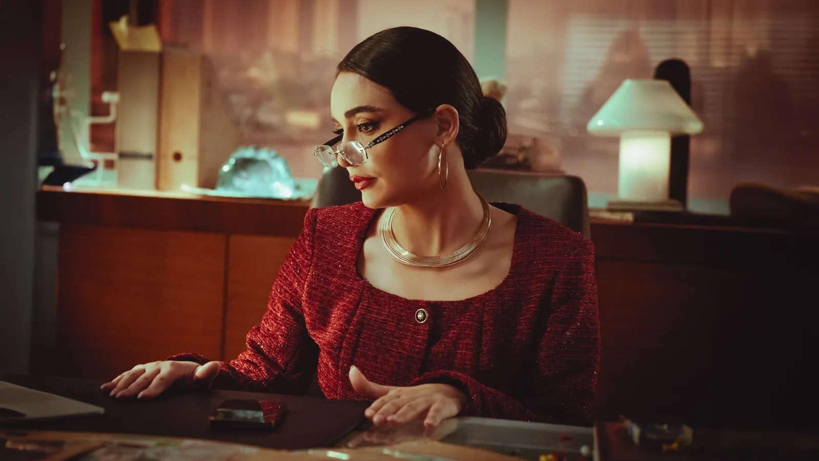 Practice owner in a burgundy tweed jacket and gold jewelry working at her desk in a warm, lamp-lit private office