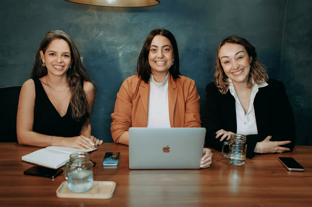 Three smiling practice leaders in blazers at a walnut conference table with a laptop, ready to meet an agency