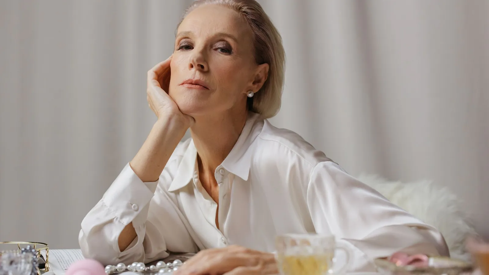 Poised silver-haired woman in a white silk blouse resting her chin on her hand at a table with pearls