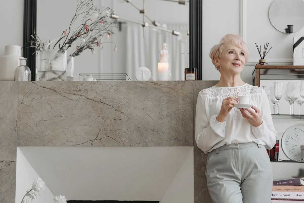 Silver-haired woman holding a cup of tea beside a stone fireplace in a refined modern home