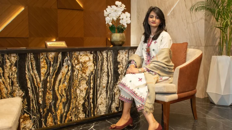Elegantly dressed woman seated beside a marble reception desk with white florals in a luxury lobby