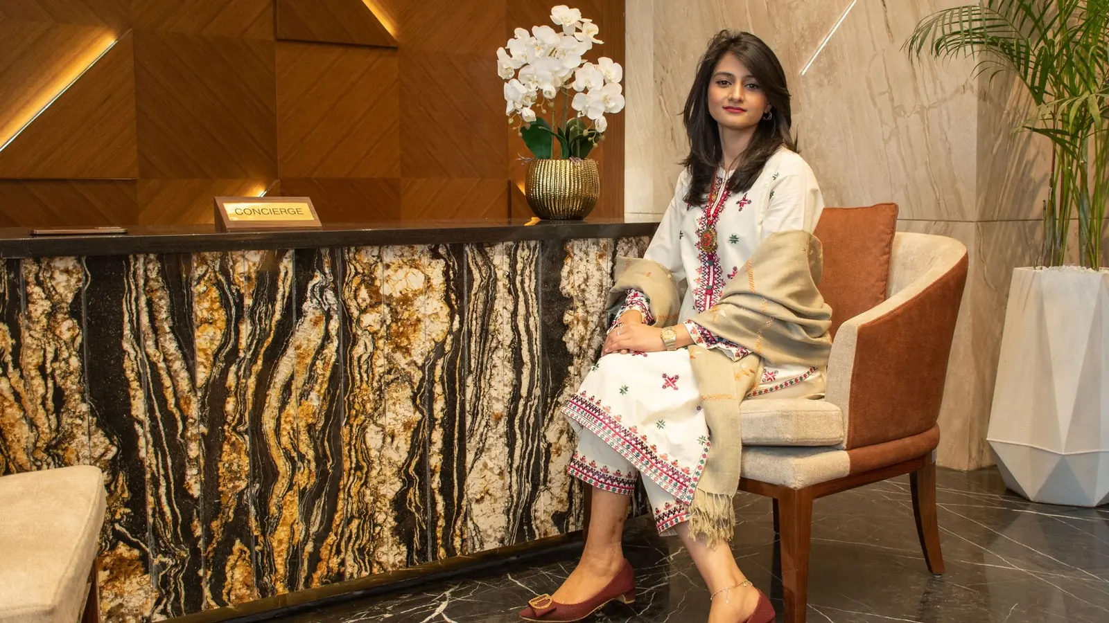 Elegantly dressed woman seated beside a marble reception desk with white florals in a luxury lobby