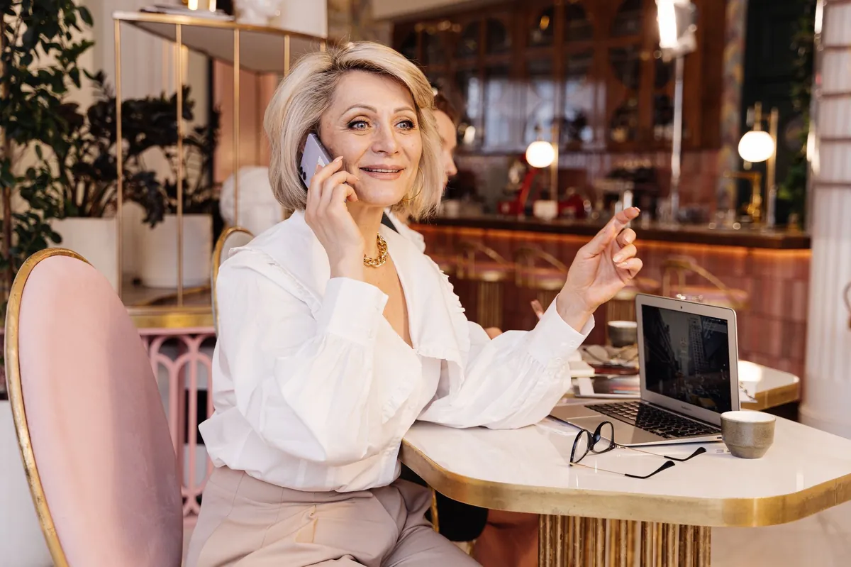 Silver-haired businesswoman on a smartphone call at a marble cafe table with her laptop