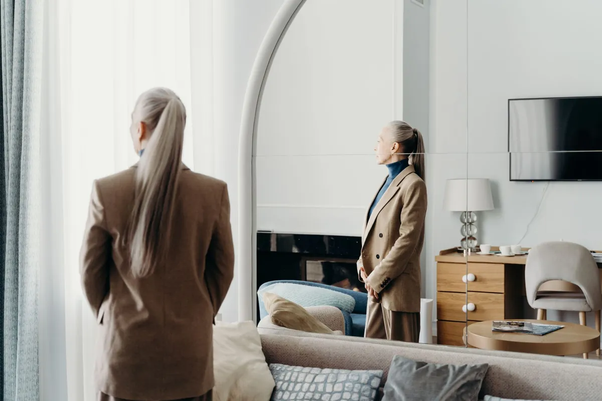Woman in a camel coat viewing her reflection in a tall mirror in an elegant room