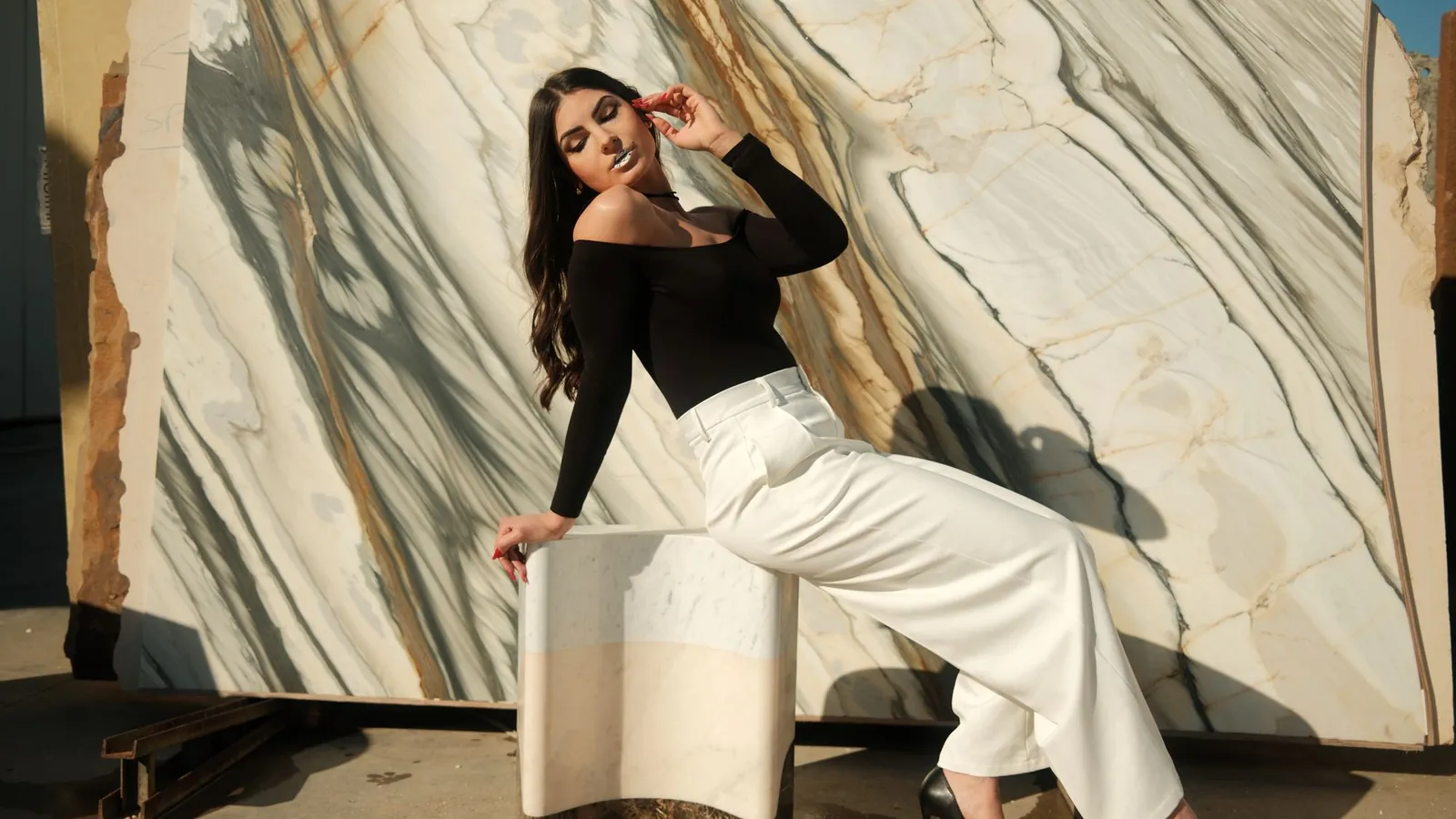 Woman in a black off-shoulder top and wide white trousers seated against a sweeping marble wall