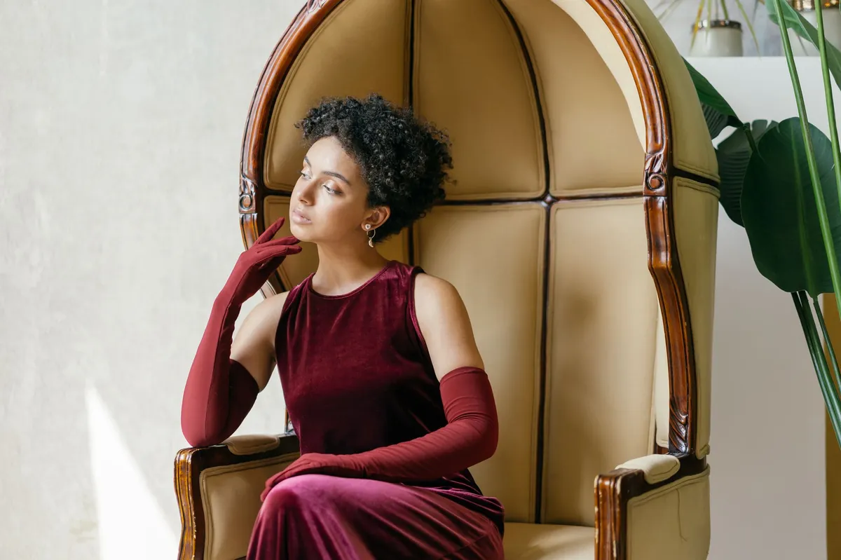 Woman in a burgundy velvet dress and opera gloves seated in an arched ivory chair