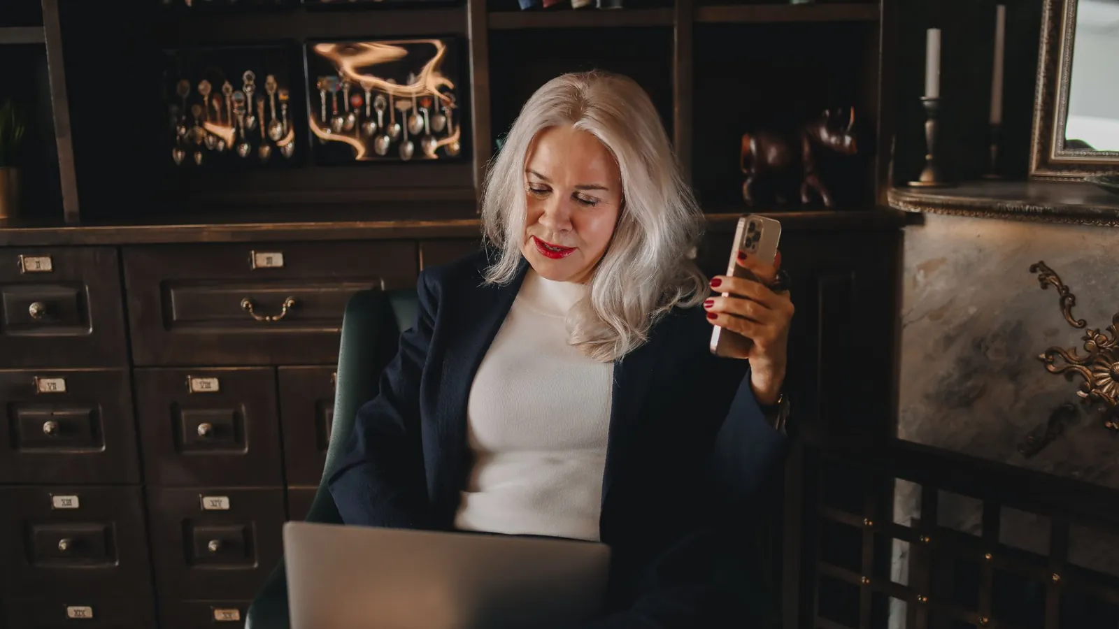 Silver-haired practice owner in a navy blazer checking her phone at a laptop in a warm, wood-paneled private office with brass details