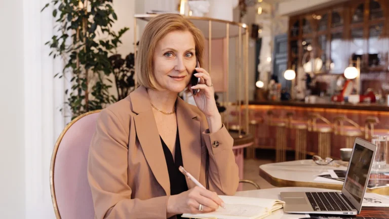 Med spa owner in a camel blazer taking a call with her laptop open at a brass-accented café table