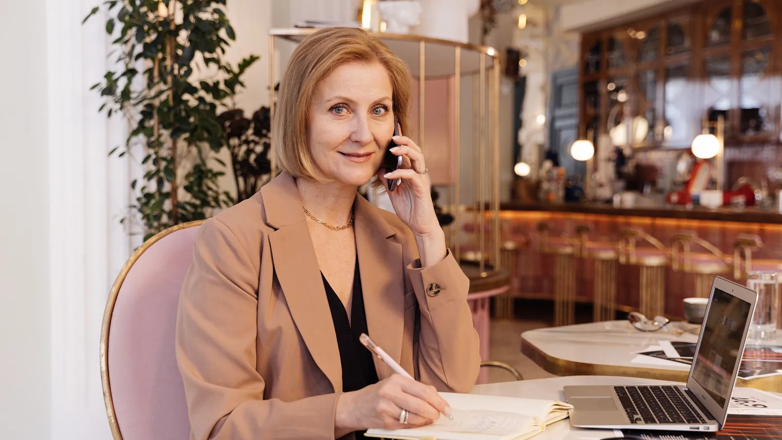 Med spa owner in a camel blazer taking a call with her laptop open at a brass-accented café table