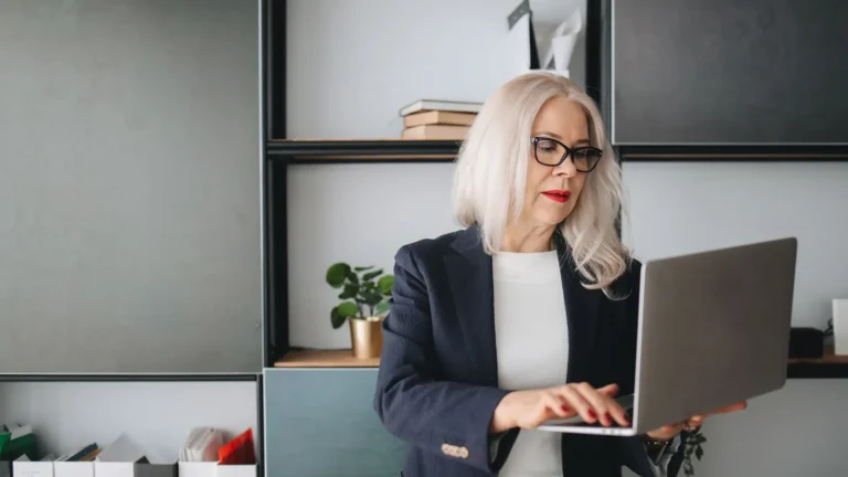 Silver-haired professional woman in a dark blazer working on a laptop in a modern office