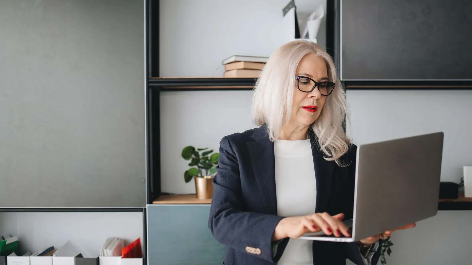 Silver-haired professional woman in a dark blazer working on a laptop in a modern office