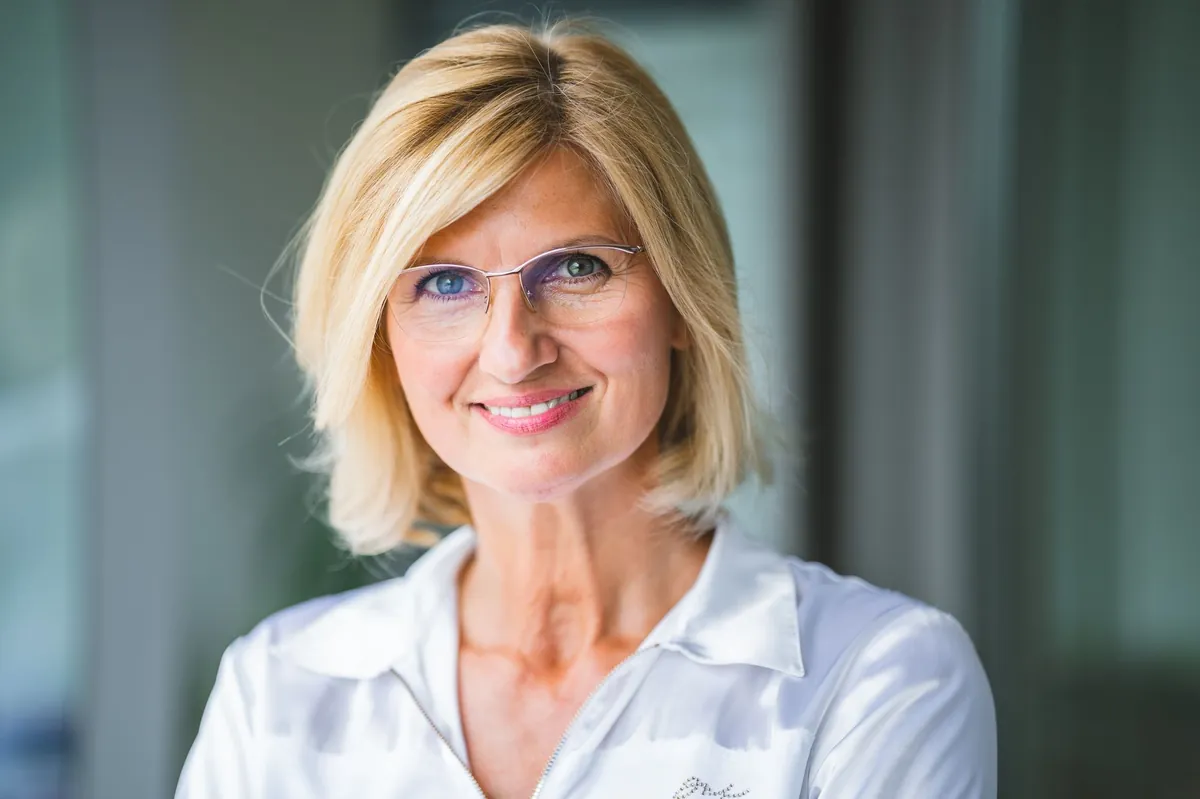 Smiling blonde woman in eyeglasses and a white blouse in a softly lit room