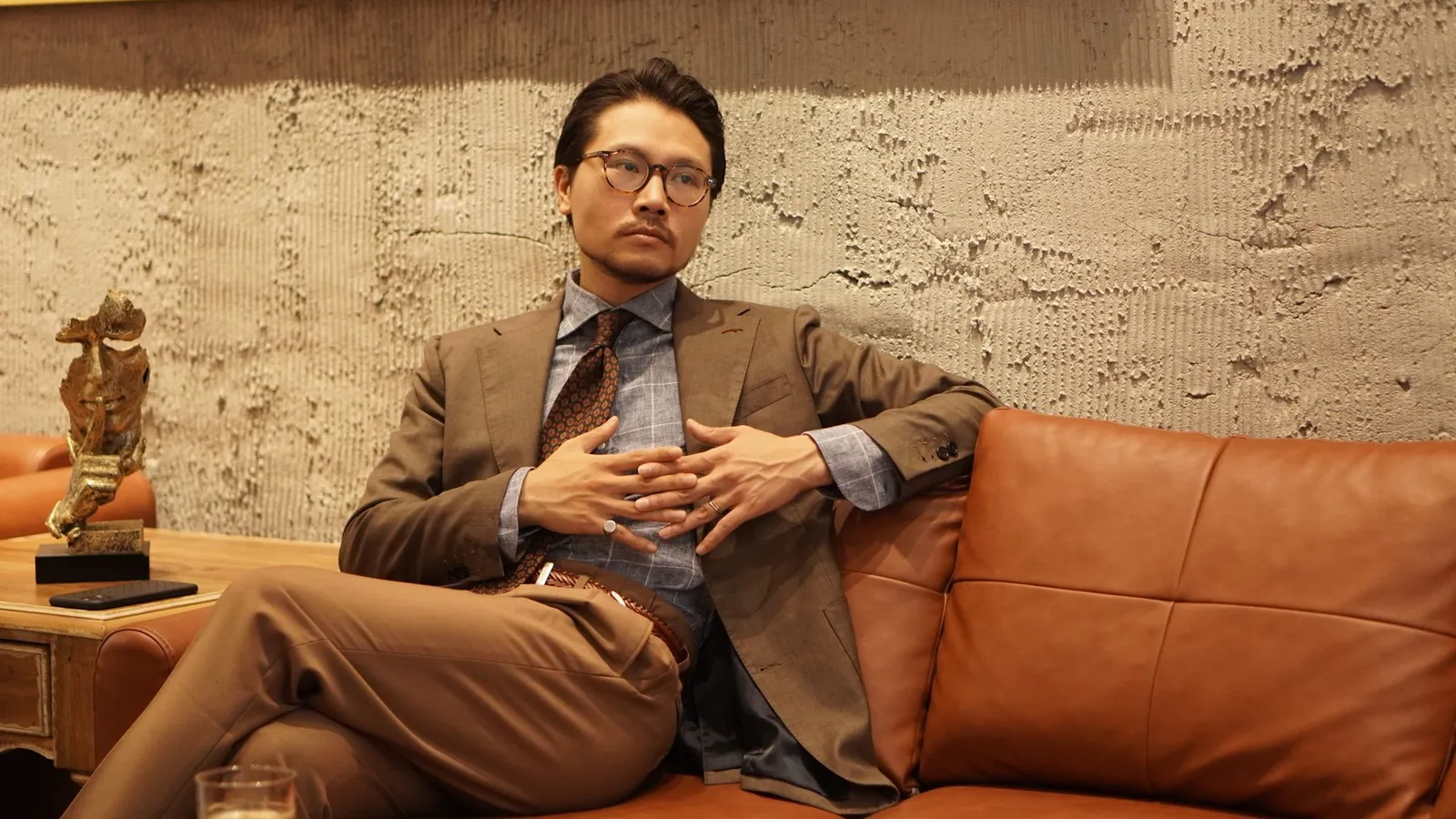 Man in a tailored brown suit and glasses seated on a cognac leather sofa