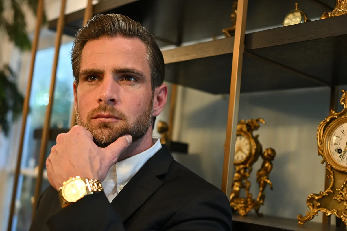 Man in a dark suit and gold watch resting his chin on his hand beside antique gilded clocks