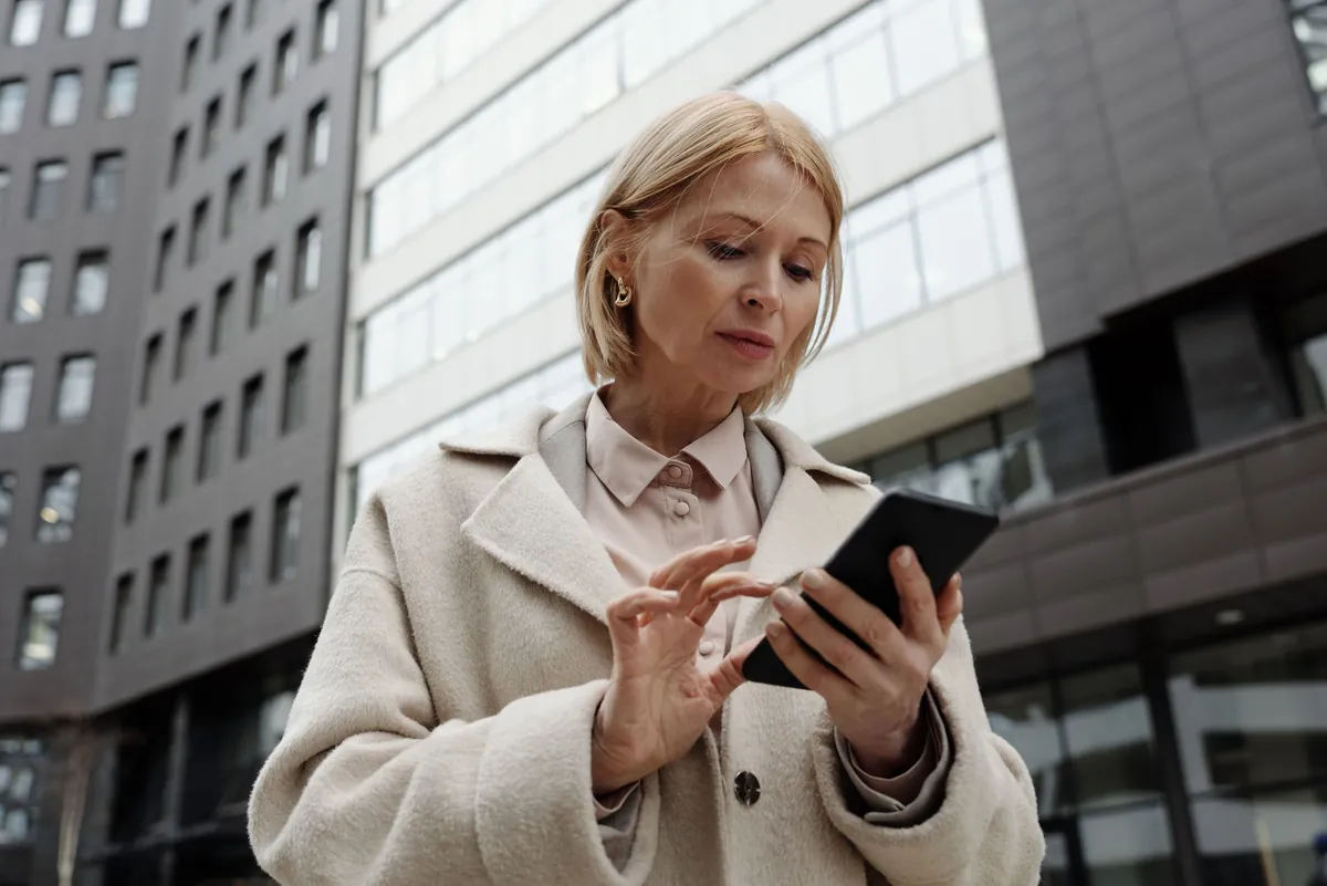 Woman in a cream coat checking her smartphone outside a modern building
