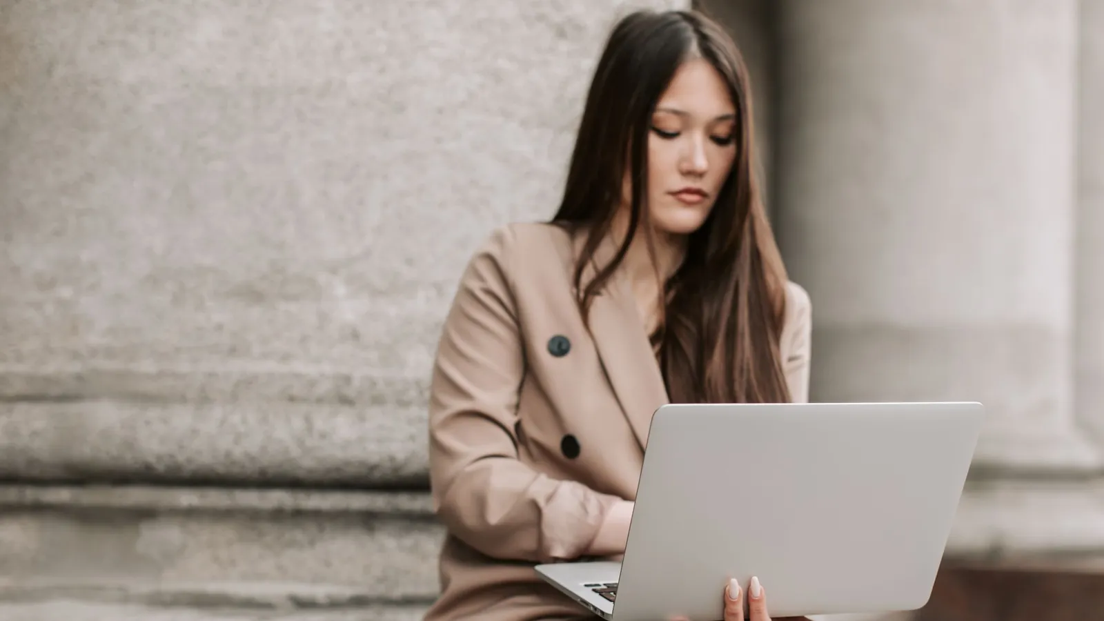 Practice owner in a beige blazer reviewing her website on a laptop beside a stone column