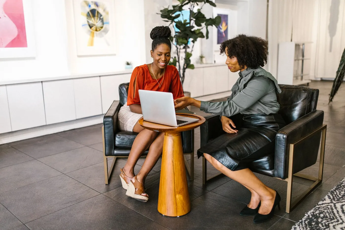Two polished practice leaders reviewing marketing results on a laptop in brass-framed leather lounge chairs in a chic gallery-style office