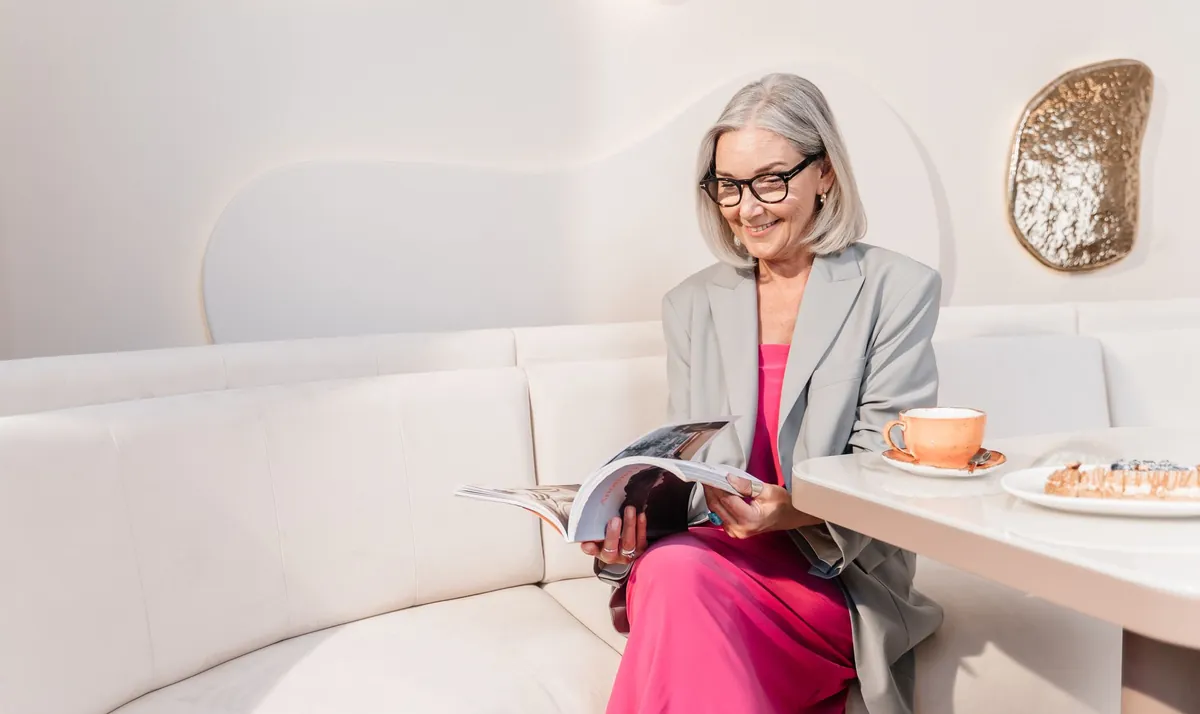 Silver-haired woman reading a magazine with coffee on a white sofa in a stylish lounge
