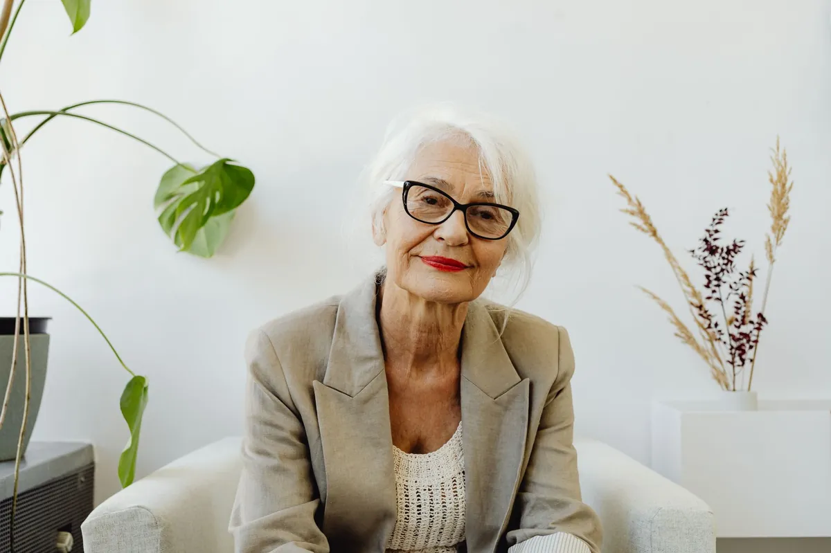 Elegant senior woman in eyeglasses and a camel blazer smiling in a bright, airy room
