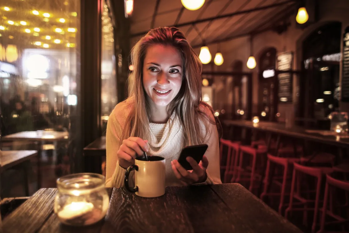 Woman smiling at her phone over coffee in a warmly lit cafe