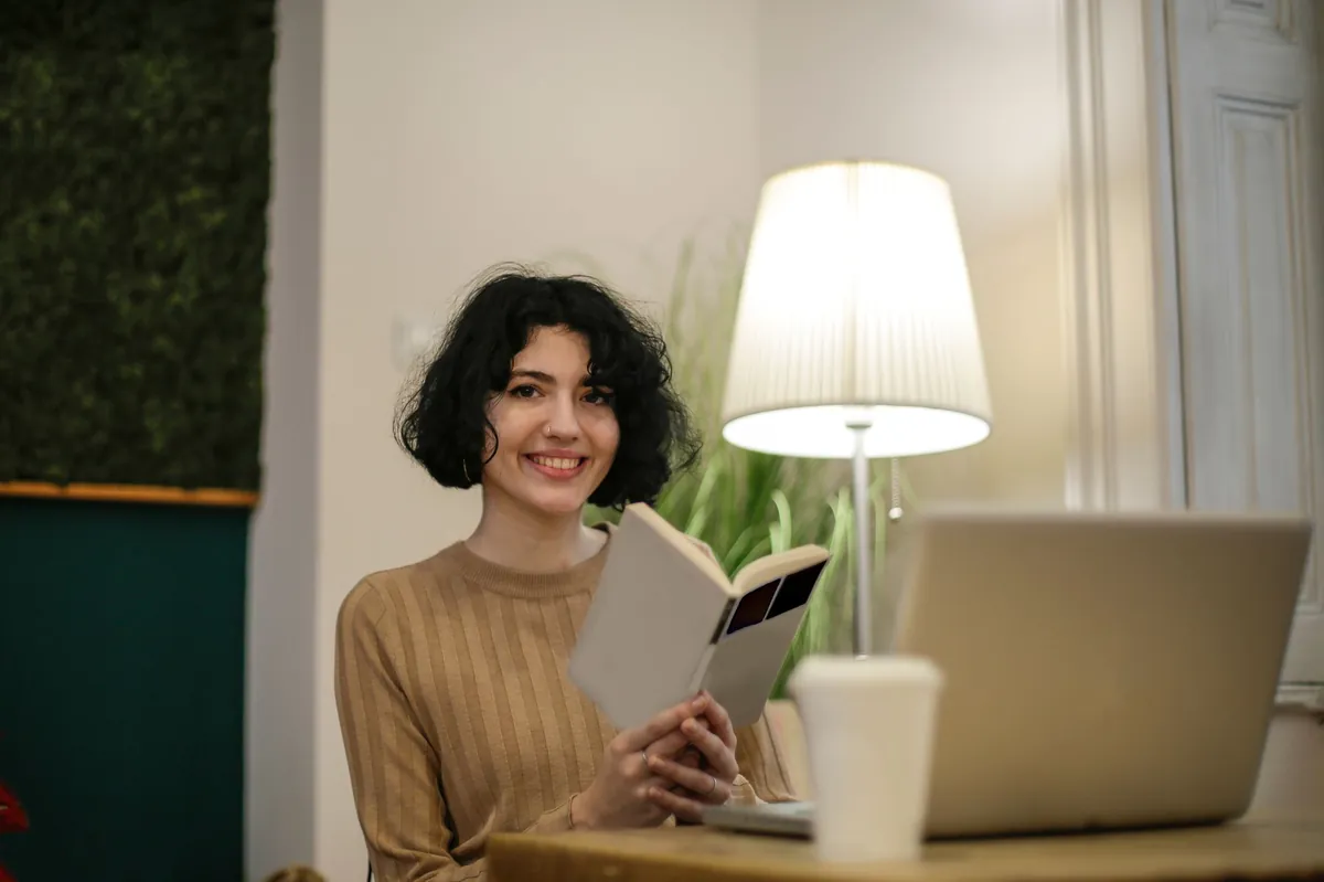 Woman with a book and laptop at a desk beside a white table lamp